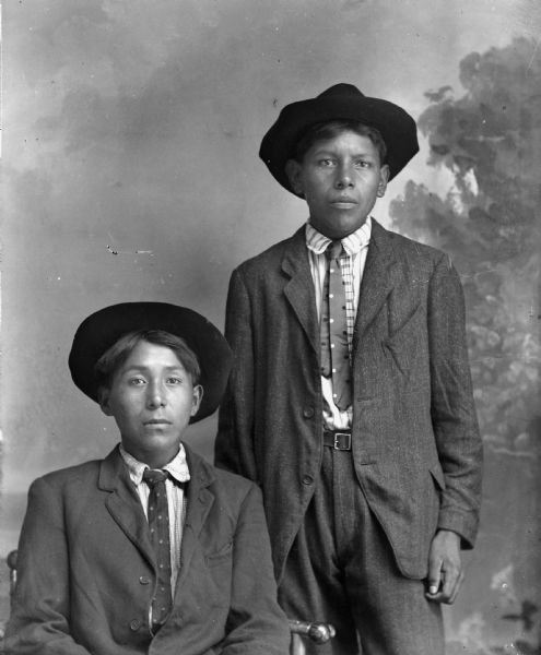 Studio portrait of a young Ho-Chunk man posing sitting on the left and a young Ho-Chunk man posing standing on the right in front of a painted backdrop. They are both wearing suits, neckties, and hats. They are identified as John Green Grass sitting on the left, and Frank Davis standing on the right.