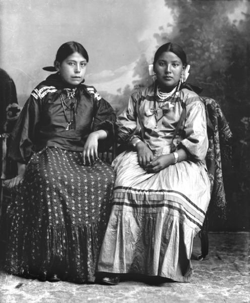 Studio portrait of two young Ho-Chunk women posing sitting in front of a painted backdrop. They are both wearing several necklaces, earrings, and ribbons in their hair. The woman sitting on the right is also wearing file bracelets and a silk ribbon banding skirt. Names, (left) Johanna Puss Otter, Greendeer, Tebo (Wa Ga Cha Me Nuk Kah) daughter of: George Otter (Mo No Ken Noo Kah) and Kate Decorra, Otter (Nau Cho Pin Win Kah) and (right) Dora Whitefeather Decorra, Greengrass (Ho Ha Win Kah) daughter of: White Feather Decorra (Ma Shu Ska) and Mary Mae Yellowbank-Beaver (Hish Chaw Skaw) (White Face Woman).