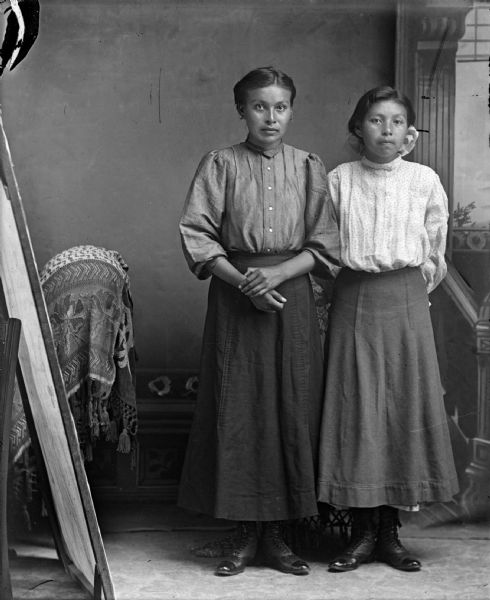 Studio full-length portrait of two young Ho-Chunk women posing standing with a light-reflecting board visible on the left. Both of the women are wearing a blouse, skirt, and boots. The woman standing on the left is identified as Jennie Young Thunder, and the woman standing on the right is identified as Lucy White Woman/Davis.