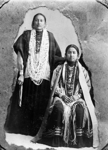 Copy photograph of a studio portrait of a Ho-Chunk woman posing standing on the left with her left hand on the right shoulder of a Ho-Chunk woman posing sitting on the right. Both are wearing regalia, including several necklaces, earrings, rings, and file bracelets. The woman on the left is wearing a wool shawl, and the woman on the right is wearing a narrow stole. The photographed image also shows cropping paper. Identified as Annie Davis standing on the left, and Little Bear Woman sitting on the right.