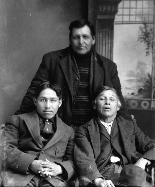 Studio portrait in front of a painted backdrop of two Ho-Chunk men sitting in front of another Ho-Chunk man standing. The younger Ho-Chunk man sitting on the left is wearing a suit and has a bandaged finger, and the elderly man sitting on the right is wearing a suit coat and is holding a hat on his lap. The man standing in the center is wearing a suit coat and striped sweater, and his face is blurred from movement. Names, left to right, Max Bearheart (Mon Ing Gah), Bert Blackspirit (Nakjaykhisepja), and Charley White (Haynaahogomikga).