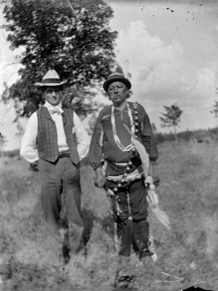 Two Ho-Chunk men posing standing in a field. The man on the left is wearing a bow tie, vest, and hat, and the man on the right is wearing a long bandoleer, several necklaces, a hat, garters and a woven sash. They are identified as (?) Johnson standing on the left and David Little Soldier standing on the right.