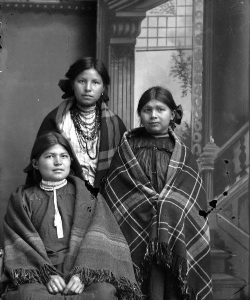 Studio portrait of a Ho-Chunk woman posing sitting on the left, and two women posing standing in the center and on the right. They are in front of a painted backdrop, and are wrapped in shawls with ribbons in their hair. The women on the left and right are wearing several short necklaces, and the woman in the center is wearing several long necklaces. They are identified from left to right as: Ethel Windblowe sitting on the left, Sarah Windblowe standing center, and Elizabeth Thunder Cloud standing on the right.