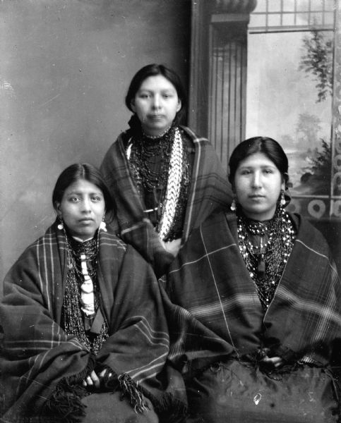 Studio portrait of two Ho-Chunk women sitting in front of another Ho-Chunk woman standing. All of the women are posing in front of a painted backdrop, and are wearing several necklaces and earrings, wrapped in plaid fringed shawls. They are identified from left to right as: Lucy Lowe Yellow Bank, Lucy White Davis, and Annie Massie/Massey Red Horn.