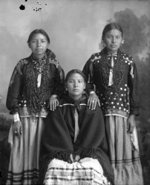 Studio portrait, in front of a painted backdrop, of a young Ho-Chunk woman posing sitting wrapped in a tied fringed shawl, between two other young Ho-Chunk women posing standing and wearing several necklaces and traditional blouses. The woman standing on the right is also wearing several earrings. They are identified from left to right as: Jenny Young Thunder, Dora Green Grass, and Lucy Davis.