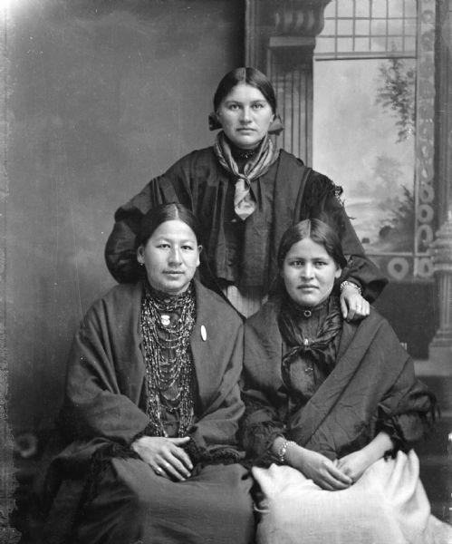 Studio portrait of two Ho-Chunk women posing sitting in front of another Ho-Chunk woman posing standing with her left arm on the left shoulder of the woman sitting on the right. The woman sitting on the left is also wearing several necklaces and a fringed shawl, and the women standing center and sitting right is wearing scarves and watches or bracelets. They are identified from left to right as: Clara St. Cyr, Lucy Davis, and Martha St. Cyr.