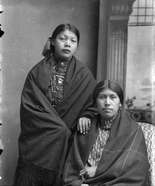Studio portrait, in front of a painted backdrop, of a Ho-Chunk woman posing standing on the left with her left arm on the right shoulder of a Ho-Chunk woman posing sitting, both wrapped in shawls and wearing necklaces. The woman standing on the left is wearing a ribbon in her hair. They are identified as Mary White Swan standing on the left, and Annie Love Lincoln sitting on the right.