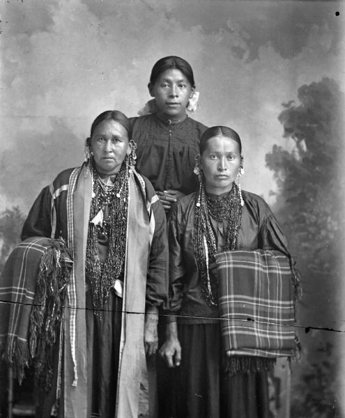 Studio portrait, taken in front of a painted backdrop, of two Ho-Chunk women posing standing in front of a Ho-Chunk woman posing standing slightly raised above the other two. The women in front are wearing several necklaces and earrings, and have folded plaid fringed shawls draped over their outside forearms. The woman in the center is wearing a dark blouse and ribbon in her hair. They are identified as Wolf Woman standing on the left, and Kate Massie standing center, woman on right unidentified.