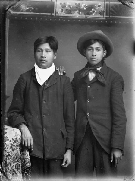 Studio portrait in front of a painted backdrop of two Ho-Chunk men posing standing. The man on the right has his right hand on the other man's left shoulder, and the man on the left has his right arm resting on a draped chair. Both men are wearing suit coats and bandanas, and the man on the right is wearing a hat. They are identified as Ed Green Grass on the left, and Frank Jefferson on the right.