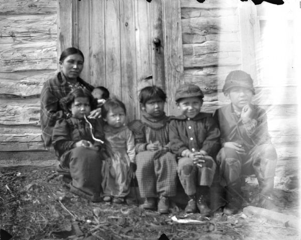 A Ho-Chunk woman and five small Ho-Chunk children and an infant are posing sitting outside the doorway of a wooden cabin. The two children on the right are wearing hats. They are identified as Myrna Lowe and her children.