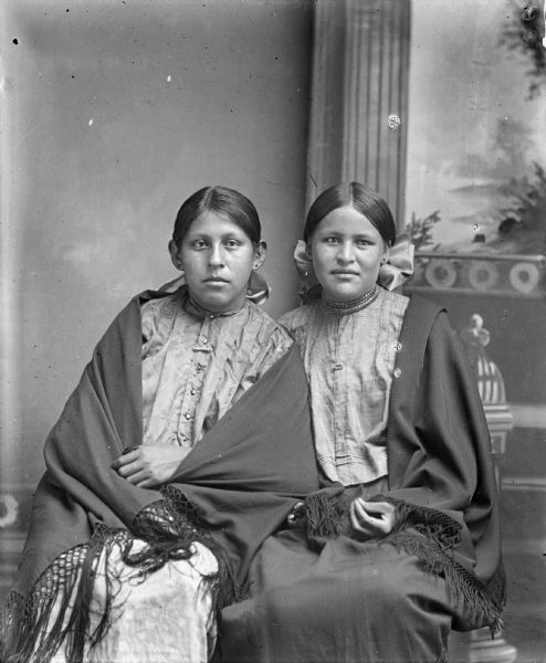 Studio portrait of two Ho-Chunk women posing sitting in front of a painted backdrop. They are wearing light-colored dresses, fringed shawls, and hair ribbons. The silk wool fringed shawls they are wearing were introduced by Peyotes from Nebraska around 1908. They are identified as Mabel White St. Cyr sitting on the left, and Minnie Stacy sitting on the right.