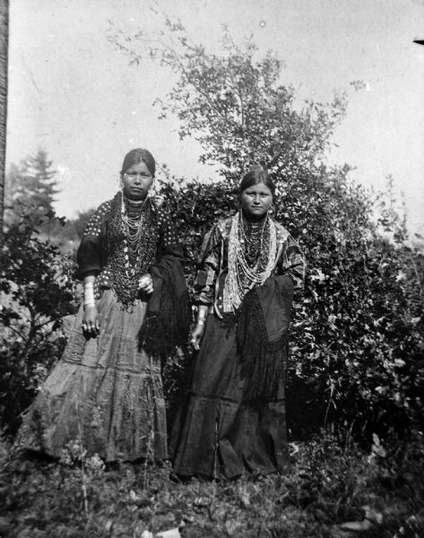 Two Ho-Chunk women outdoors posing standing in a field in front of some bushes. They are wearing long dresses, several necklaces, file bracelets, and traditional blouses, and are holding fringed shawls draped over their left forearms.