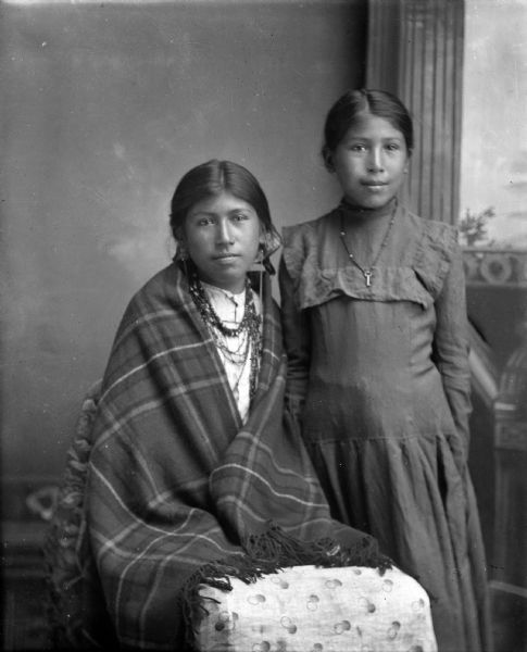 Studio portrait of a Ho-Chunk girl posing sitting in front of a painted backdrop on the left, wearing several necklaces, earrings, and wrapped in a plaid shawl. A Ho-Chunk girl is posing standing on the right, wearing a dark dress and a necklace with a small key.