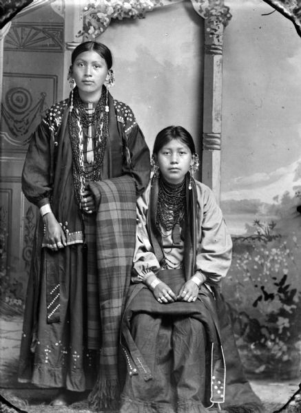 Studio portrait in front of a painted backdrop of a young Ho-Chunk woman standing on the left holding a shawl draped over her left forearm, and another Ho-Chunk girl sitting next to her with a shawl draped over her lap. Both women are wearing several necklaces, earrings, rings, file bracelets, and traditional blouses.