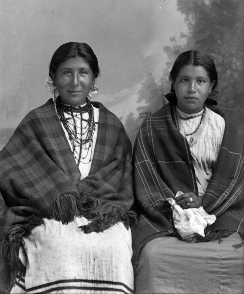 Studio portrait of two Ho-Chunk women sitting in front of a painted backdrop. They are wearing fringed plaid shawls draped over their shoulders, ribbons in their hair, and necklaces. The woman sitting on the left is wearing earrings, and the woman sitting on the right is holding a white handkerchief on her lap in her left hand.