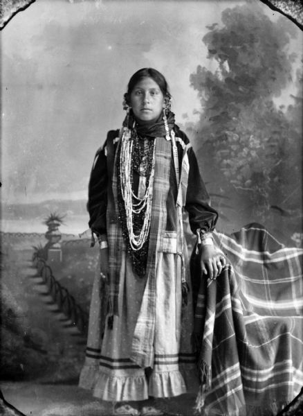 Studio full-length portrait of a Ho-Chunk girl posing standing in front of a painted backdrop. Her left hand is resting on a draped chair and she is wearing several necklaces, rings, earrings, a plaid stole, and file bracelets.