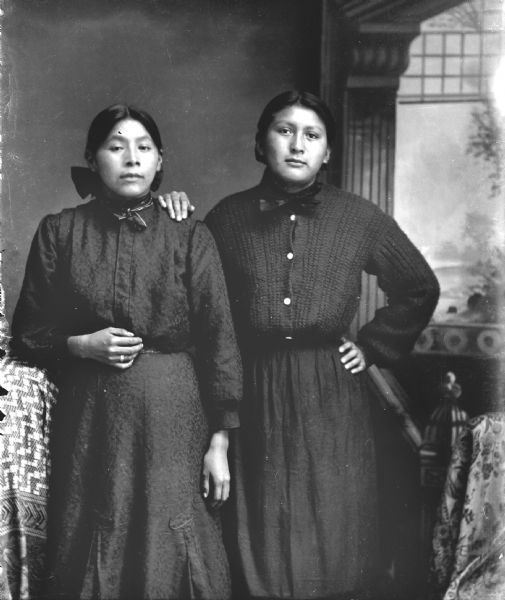Studio portrait of two Ho-Chunk women posing standing in front of a painted backdrop. On the left is Kate Massey, daughter of William Massey and Jane Winneshiek. Kate was the first cousin to George Lonetree. Both women attended Indian school in Toldo, Iowa. The woman on the right has her right hand on the other woman's left shoulder, and the woman on the left is wearing a ring and a bow in her hair. Both women are wearing dark-colored dresses with ties at the neck.