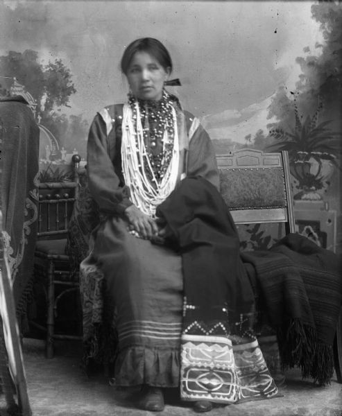 Studio portrait of a Ho-Chunk woman posing sitting in front of a painted backdrop. There are chairs and a blanket around her. She is wearing several necklaces, earrings, and a hair ribbon, and is holding a ribbon-work shawl draped over her left forearm and lap.