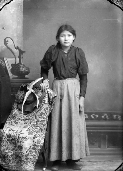 Studio full-length portrait of a young Ho-Chunk woman posing in front of a painted backdrop. She is standing with her right hand on a feathered hat resting on a chair draped with a floral throw. She is wearing a dark-colored blouse and light-colored skirt.