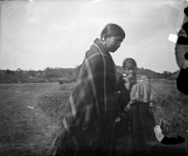 A woman and two Ho-Chunk girls are standing outdoors in a field. The woman on the left is wrapped in a shawl. The face of a girl can be seen just behind her. Another girl is standing behind the other two. In the far background is a low hill.
