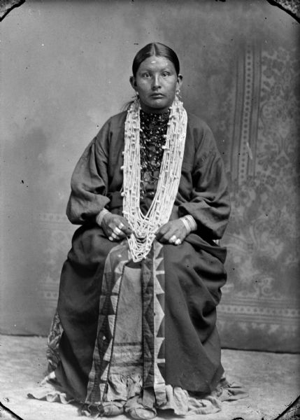Studio portrait of a Ho-Chunk woman posing sitting in front of a painted backdrop. She is wearing several necklaces, earrings, rings, file bracelets, moccasins, and a ribbon-work skirt.