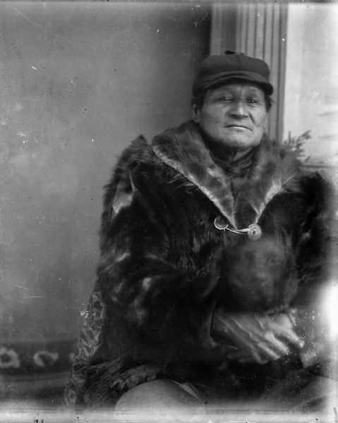 Waist-up studio portrait of an older Ho-Chunk man posing sitting in front of a painted backdrop. He is holding a dog (blurred by movement) in his lap, and is wearing a fur coat and hunting hat.