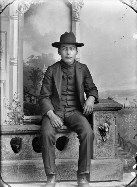 Studio portrait of a young Ho-Chunk man, George Otter, Jr. (Hay Cho Kah), sitting on a prop stone wall in front of a painted backdrop. He is wearing a suit coat, vest, bandana, and hat.