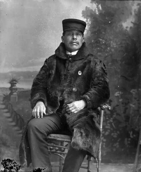 Studio portrait of a Ho-Chunk man posing sitting in front of a painted backdrop. He is wearing a long fur coat and a Scotch cap.