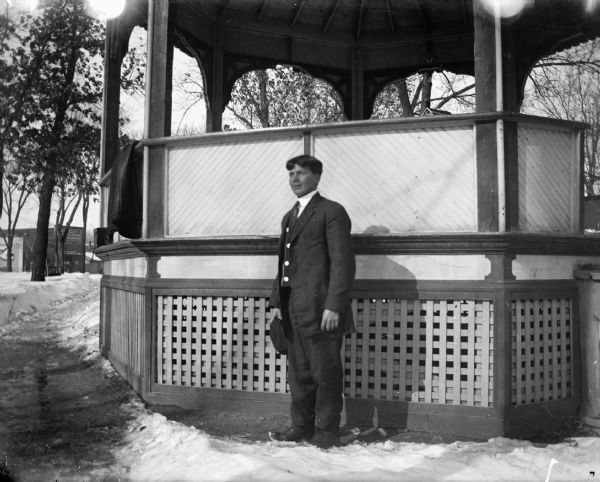A man is posed standing on snow-covered ground in front of a gazebo in a park. He is wearing a suit and necktie, and holding a hat in his right hand. There is a coat draped over the railing of the gazebo on the left. In the background are trees and storefronts.
