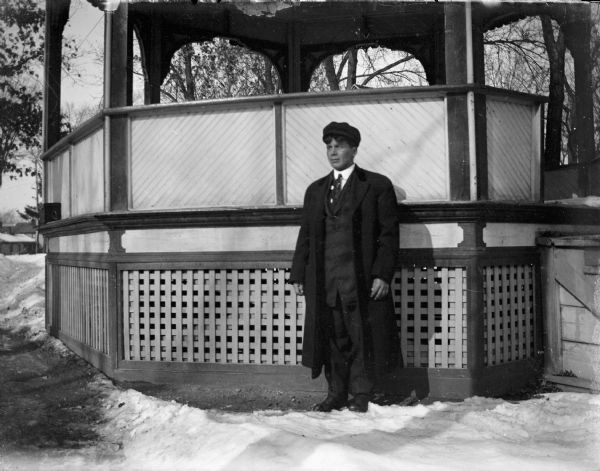 A man is posed standing on snow-covered ground in front of a gazebo in a park. He is wearing a coat, suit and necktie, and holding a hat in his right hand. In the background are trees and buildings.