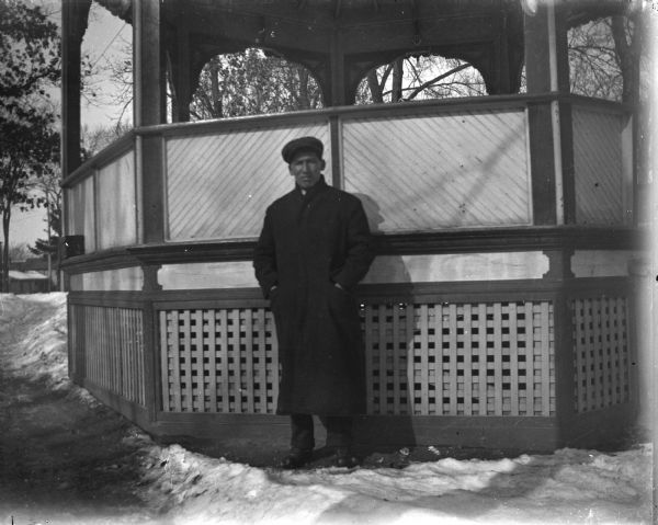 Ho-Chunk man posing standing on the snow-covered ground in front of a gazebo. He is wearing an overcoat, necktie, and hat. There are trees in the background, and buildings on the left side.