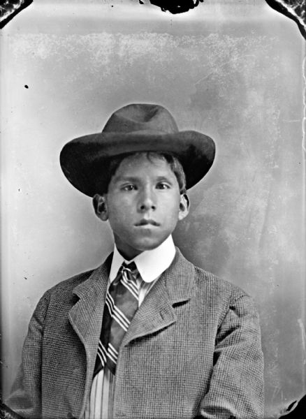 Studio portrait of a Ho-Chunk boy posing sitting and wearing a suit coat, slip tie, and hat.
