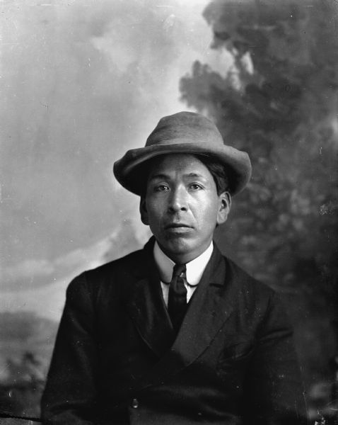 Waist-up studio portrait of a Ho-Chunk man sitting in front of a painted backdrop. His name is Frisk Cloud (Ho-Chunk from the Tomah area). He is wearing a suit coat, necktie, and hat.