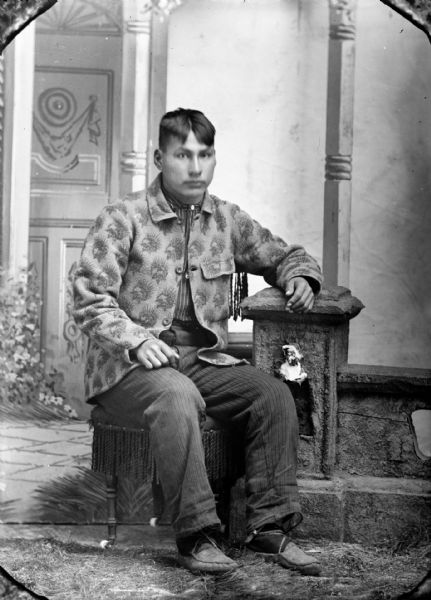 Studio portrait of a young Ho-Chunk man posing sitting on a stool in front of a painted backdrop. He has his left arm resting on a prop stone wall, and is wearing a dark-colored striped shirt and Indian-head print jacket.