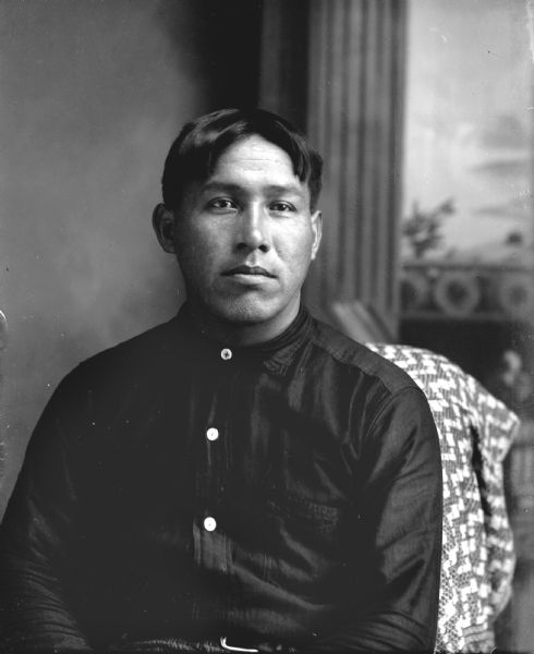 Waist-up studio portrait of Max Bearheart (Mon Ing Gah) with short hair sitting in front of a painted backdrop. He is wearing a dark button-up shirt.