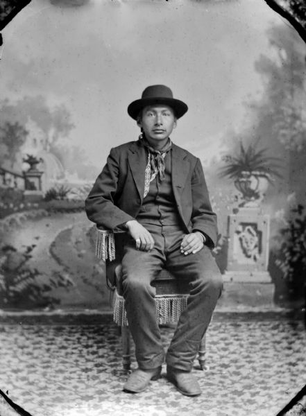Full-length studio portrait of a Ho-Chunk man posing sitting in a tasseled chair in front of a painted backdrop. He is wearing a suit, bandana, hat, and a ring on the pinky finger of his right hand.