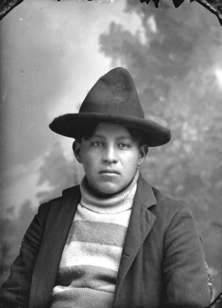 Quarter-length studio portrait of a young Ho-Chunk man posing sitting in front of a painted backdrop. He is wearing a striped turtleneck sweater, suit coat, and hat.