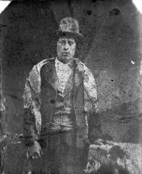 Copy photograph of a poor quality studio portrait of a Ho-Chunk man posing standing. He is wearing a suit, hat, and shawl over his shoulder. This is possibly originally a tintype.