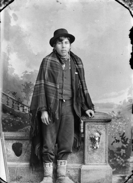 Full-length studio portrait of John Pigeon – “He Cho Kah.” Born 1875, died August 1915. Posing standing in front of a prop stone wall and painted backdrop. He is wearing a suit, bandana, earrings, garters, felt leggings, moccasins, and a Kenosha Mill shawl over his shoulders.