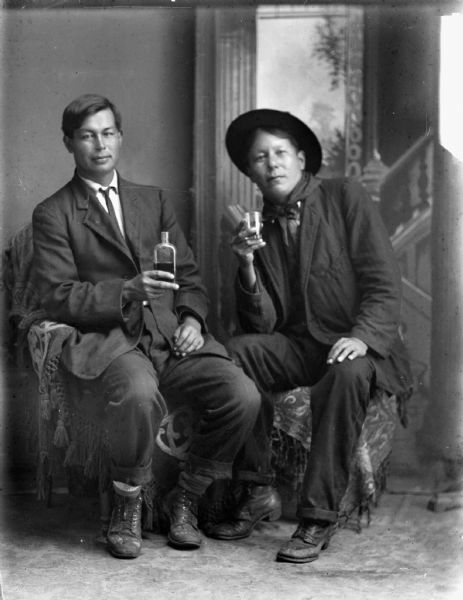 Studio portrait of two Ho-Chunk men posing sitting in front of a painted backdrop. The man on the left is holding a liquor bottle, and the man on the right is holding a small glass. The man on the left is wearing a suit coat and necktie, and the man on the right is wearing a suit coat, bandana, and hat.