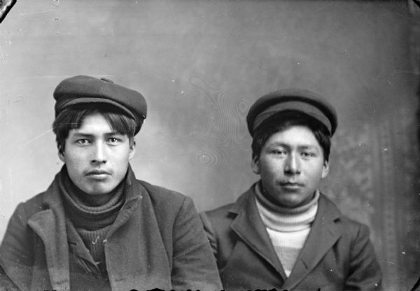 Quarter-length studio portrait of a two young Ho-Chunk men posing sitting. They are wearing suit coats, turtleneck sweaters, and caps.