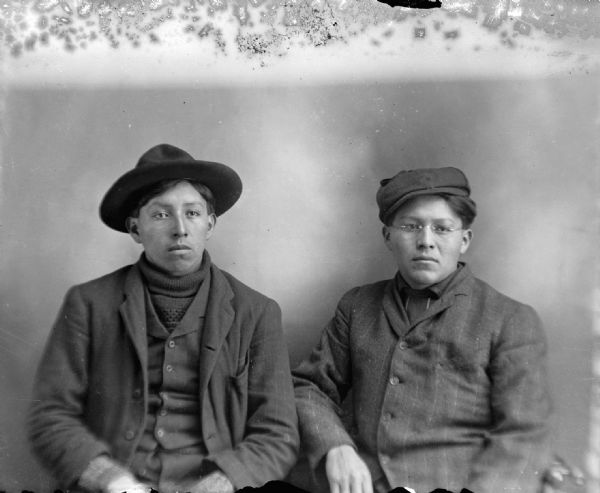 Waist-up studio portrait of two Ho-Chunk men posing sitting. They are wearing suit coats and vests, the man on the left is wearing a hat and turtleneck sweater, and the man on the right is wearing a cap and eyeglasses.