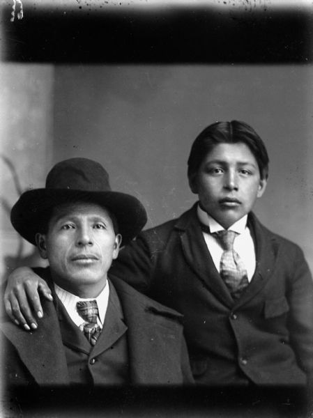 Studio portrait of two Ho-Chunk men posed seated. They are wearing suit coats and neckties. The younger man on the right has his arm around the shoulders of Fred Kingswan, on the left, who is wearing an overcoat and hat.