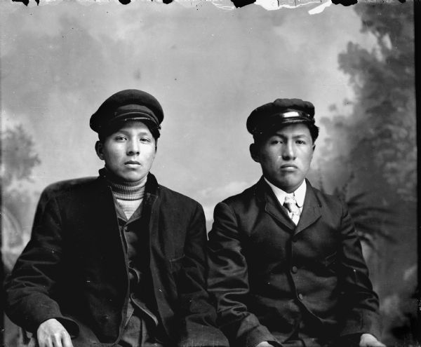 Waist-up studio portrait of two young Ho-Chunk men posing sitting in front of a painted backdrop wearing suit coats and caps. The man on the left is wearing a turtleneck sweater, and the man on the right is wearing a necktie.