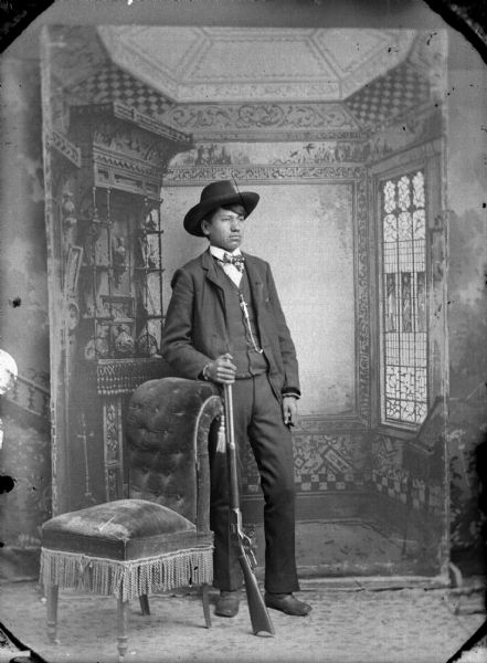Full-length studio portrait of a young Ho-Chunk man standing next to a fringed, plush chair. He is holding a rifle with his right hand, and is wearing a suit, bow tie, pocket watch chain, and hat. He is identified as Thomas Thunder (Hoonk Ha Ga Kah) (Son of [Wa Con Cha Kah] John Thunder aka Dr. Thunder and [We Hon Pe Kaw] Lucy Bear, Thunder). In the background is a painted backdrop.