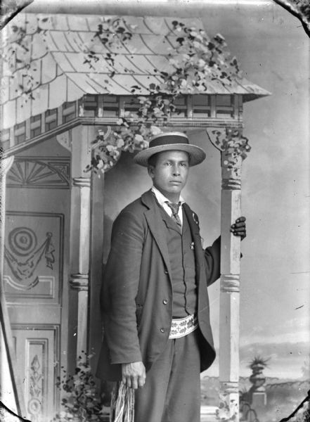 Three-quarter length studio portrait of a Ho-Chunk man posing standing in front of a painted backdrop. He is holding a prop pillar with his left hand, and is wearing a suit, necktie, woven belt, and straw hat.