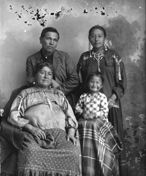 Studio portrait of a Ho-Chunk group. Rachel Funmaker, Winneshiek is sitting on the left wearing earrings, and a small child is on the right sitting on a stool with a shawl over his legs, and wearing a plaid shirt and gorget. They are seated in front of a man who is standing on the left and wearing a suit coat, and Grace Waukon, Green who is standing on the right wearing several necklaces and earrings. In the background is a painted backdrop.