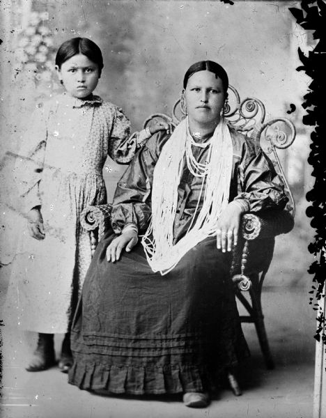 Studio portrait in front of a painted backdrop of a Ho-Chunk girl posing standing on the left wearing a light-colored print dress, and a Ho-Chunk woman posing sitting on a elaborate wicker chair on the right, wearing several necklaces, earrings, file bracelets, and dark-colored dress.