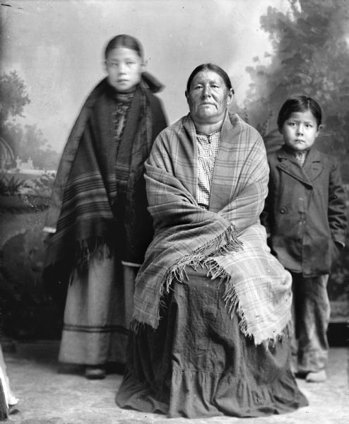Studio portrait of Mary Snowball, Decorra, Buffalohead (Pay Dok Se Kah), (Daughter of [Wau Soo Mon E Kah] Snow Ball and mother [Wa Con Cha Ka Win Kah], she is also the maternal granddaughter of Iron Walker and Green Feather Woman). She is wrapped in a fringed plaid shawl, and a Ho-Chunk girl and boy are on either side of her. The girl is standing on the left wrapped in a fringed striped shawl, and the boy with short hair is standing on the right wearing a suit coat. In the background is a painted backdrop.