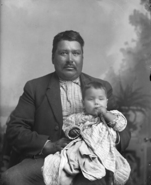 Studio portrait of Jim White Bear posed sitting in front of a painted backdrop wearing a suit coat. In his lap he is holding a Ho-Chunk infant wearing a light-colored dress.